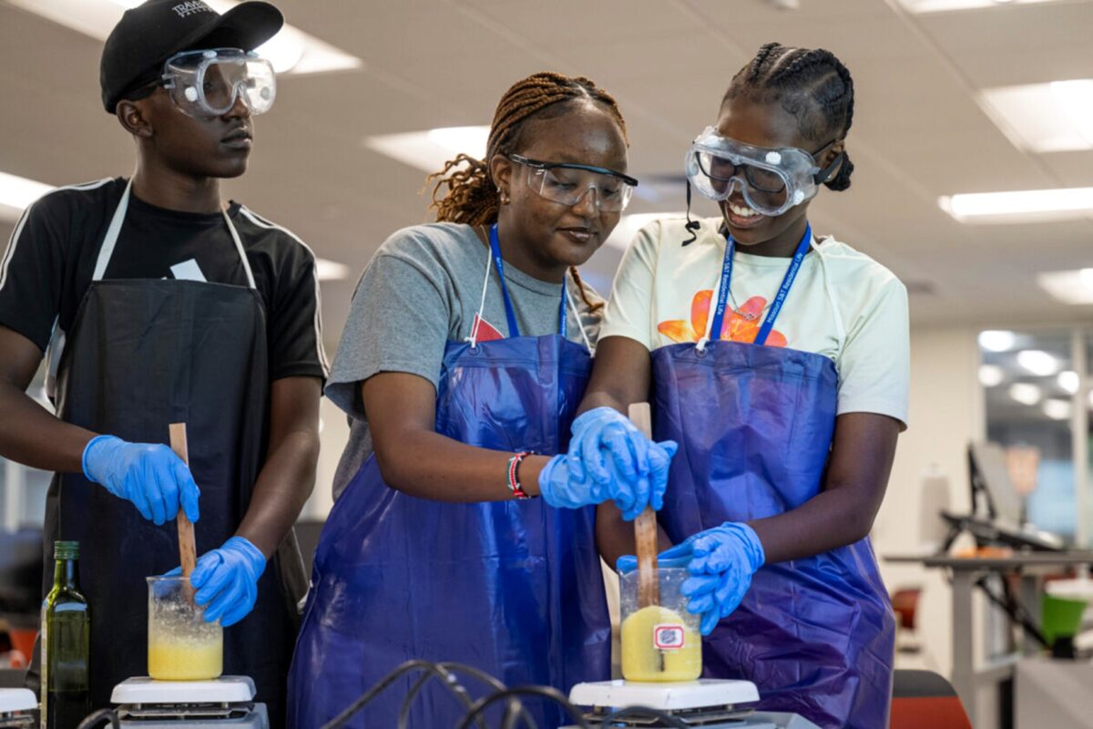 Three summer campers mix a substance in a beaker while wearing gloves, aprons and safety goggles.