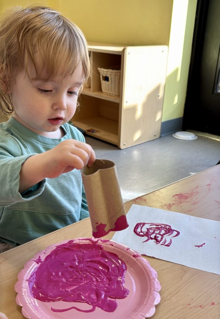 A toddler uses a toilet paper tube shaped like a heart to stamp heart shapes in pink paint on a piece of paper.