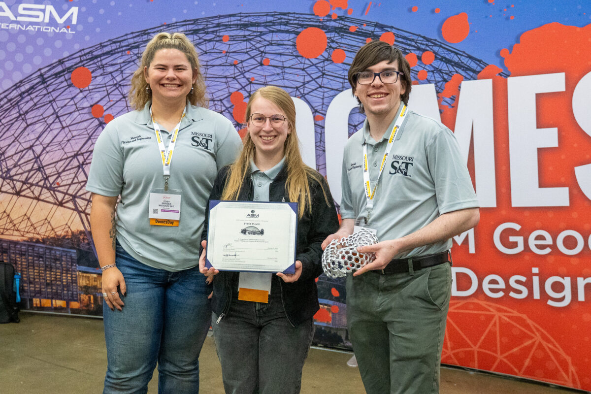 Three students wearing lanyards with one person holding a certificate and another holding the geodesic dome they submitted for the competition.