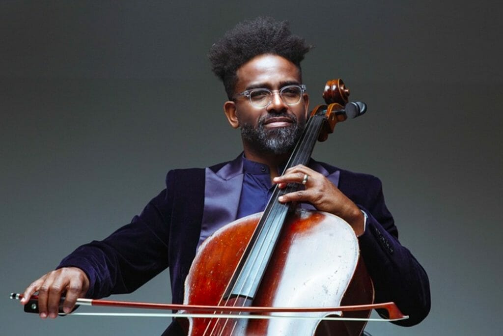 Man with glasses plays a cello, wearing a dark velvet jacket against a neutral background. His expression is focused and calm, conveying passion for music.