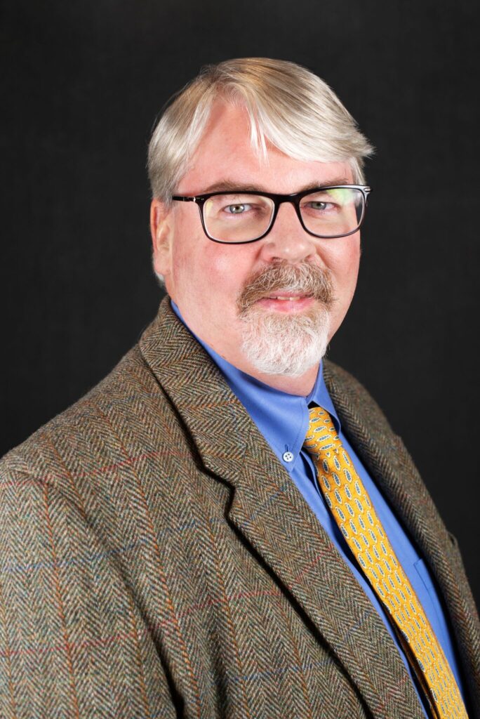 A professional headshot of Dr. Michael Bruening wearing a brown jacket, blue shirt and yellow tie. 