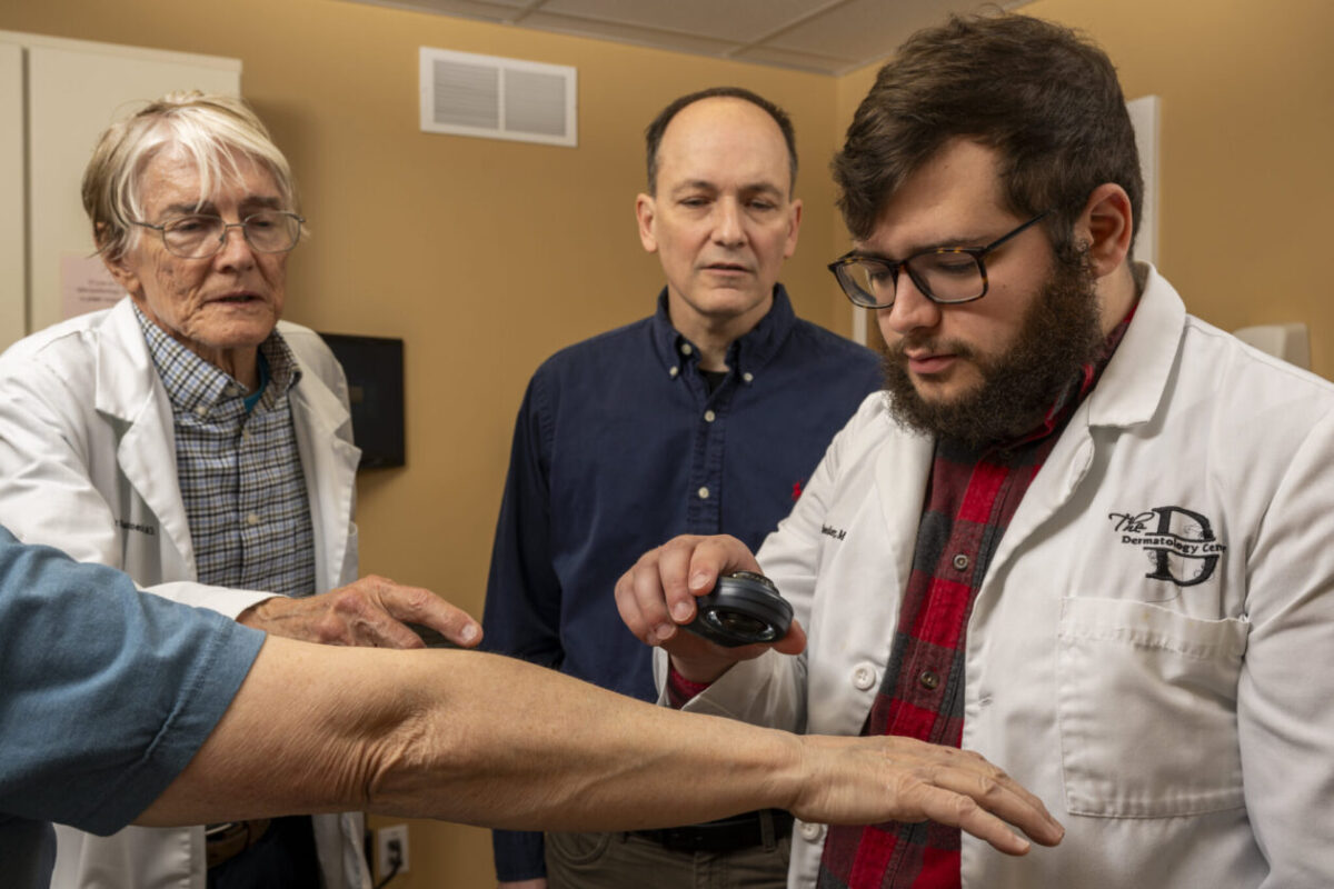 A bearded man in a white coat examines a patient's arm with a dermoscope, while two individuals observe attentively in a clinical setting.