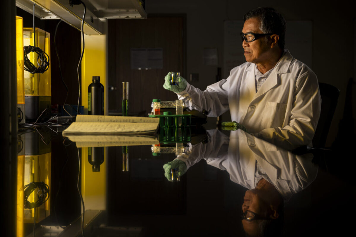 A scientist wearing a lab coat and gloves handles lab equipment at a workstation with a notebook, glass containers, and yellow-lit devices.