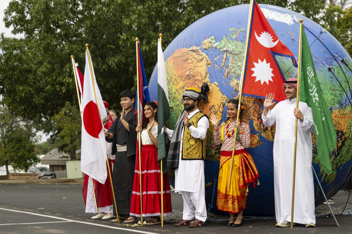 A group of people in traditional attire stand holding different national flags in front of a large globe. The mood is festive and diverse.