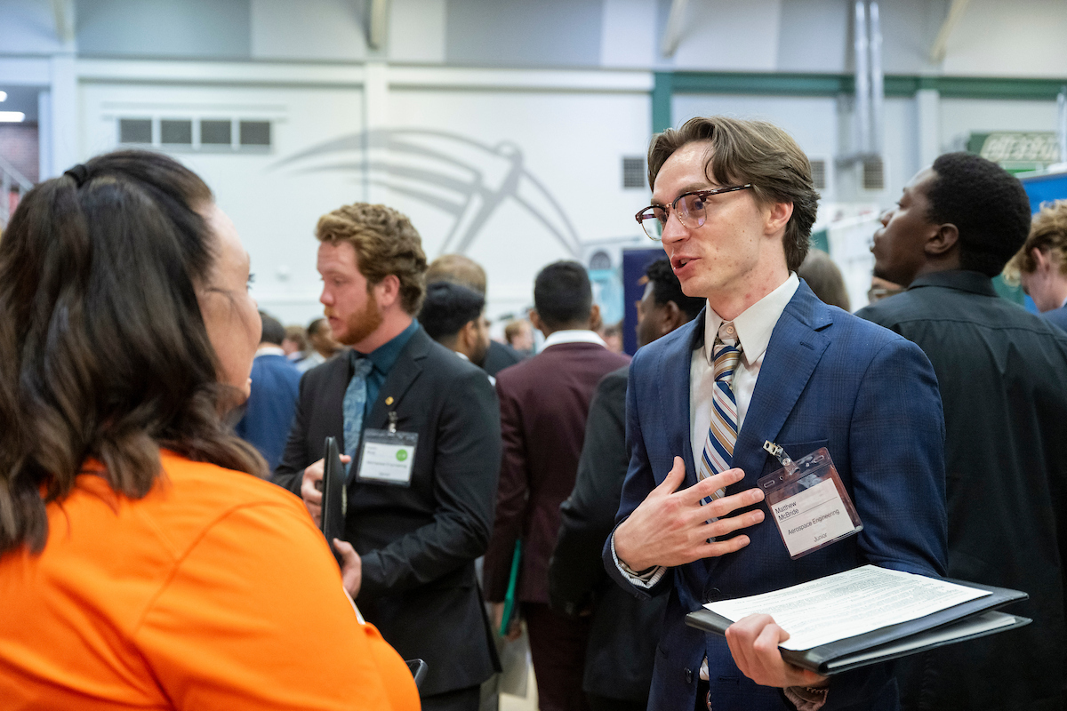 A man in a blue suit engages in conversation with a woman in an orange shirt at a crowded career fair. He holds a resume, conveying professionalism and focus.