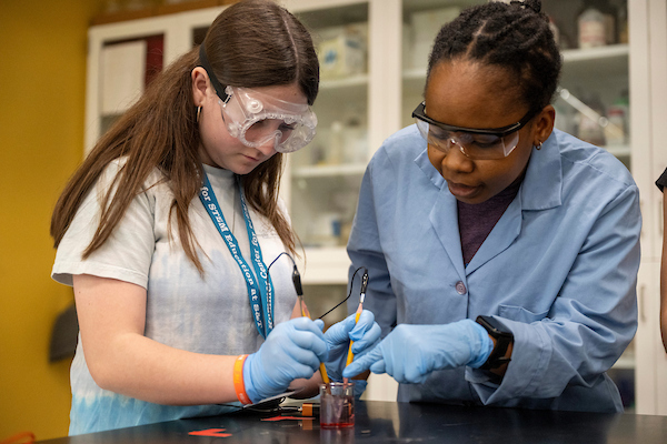 Two people in a lab conduct an experiment, wearing gloves and goggles. One guides the other in handling a tool, showing focus and collaboration.