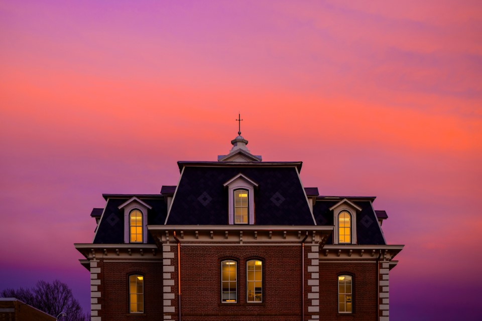 Historic brick building with a mansard roof lit from within, set against a vibrant purple and pink sunset sky. Warm, serene atmosphere.
