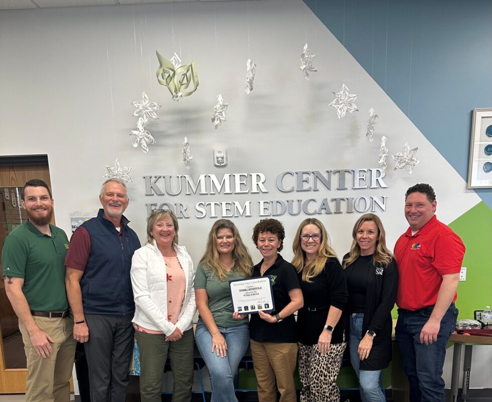 A group of eight smiling adults stands in front of the "Kummer Center for STEM Education" sign. One person holds a certificate. The atmosphere is joyful.