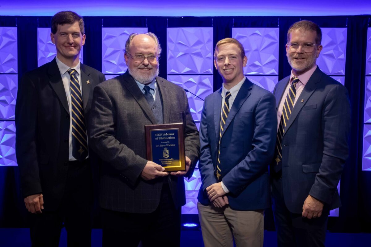 Four men wearing business attire pose in front of a purple wall with one holding a plaque.