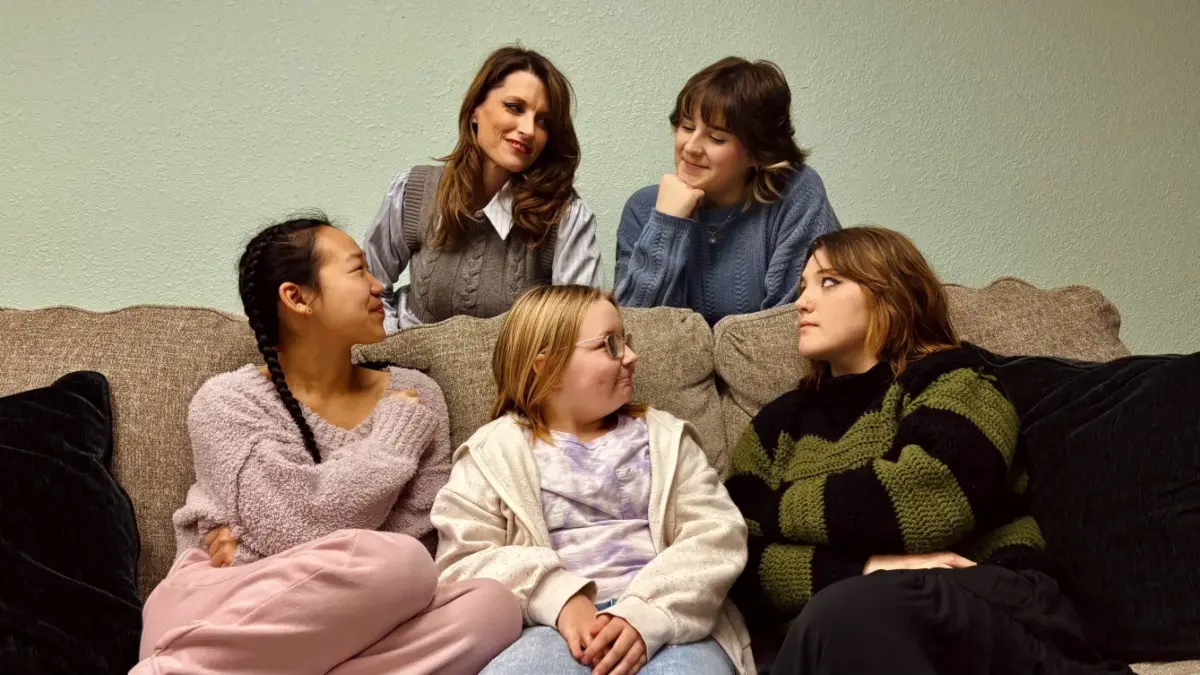 Three young women are sitting on a couch with two women standing behind them. They all are looking at each other.
