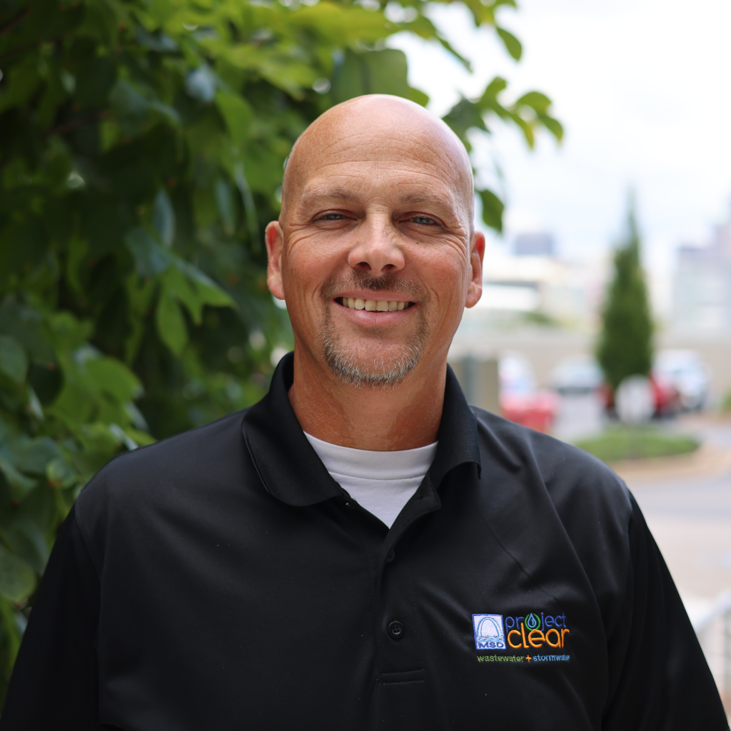 Man smiling in a black "Project Clear" polo shirt stands outdoors near green foliage, conveying a friendly and approachable vibe.