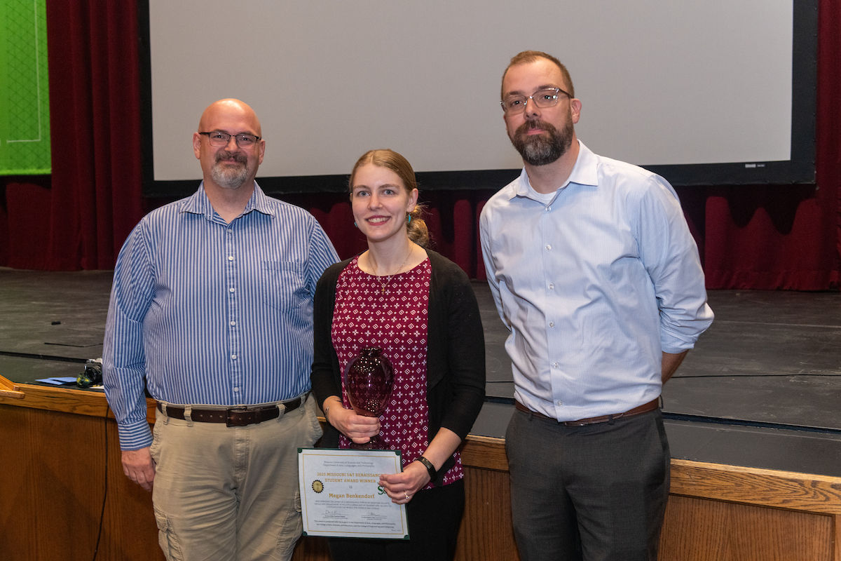 Three adults pose indoors on a stage; the woman in the center holds a plaque and a certificate, while the two men stand on either side of her.