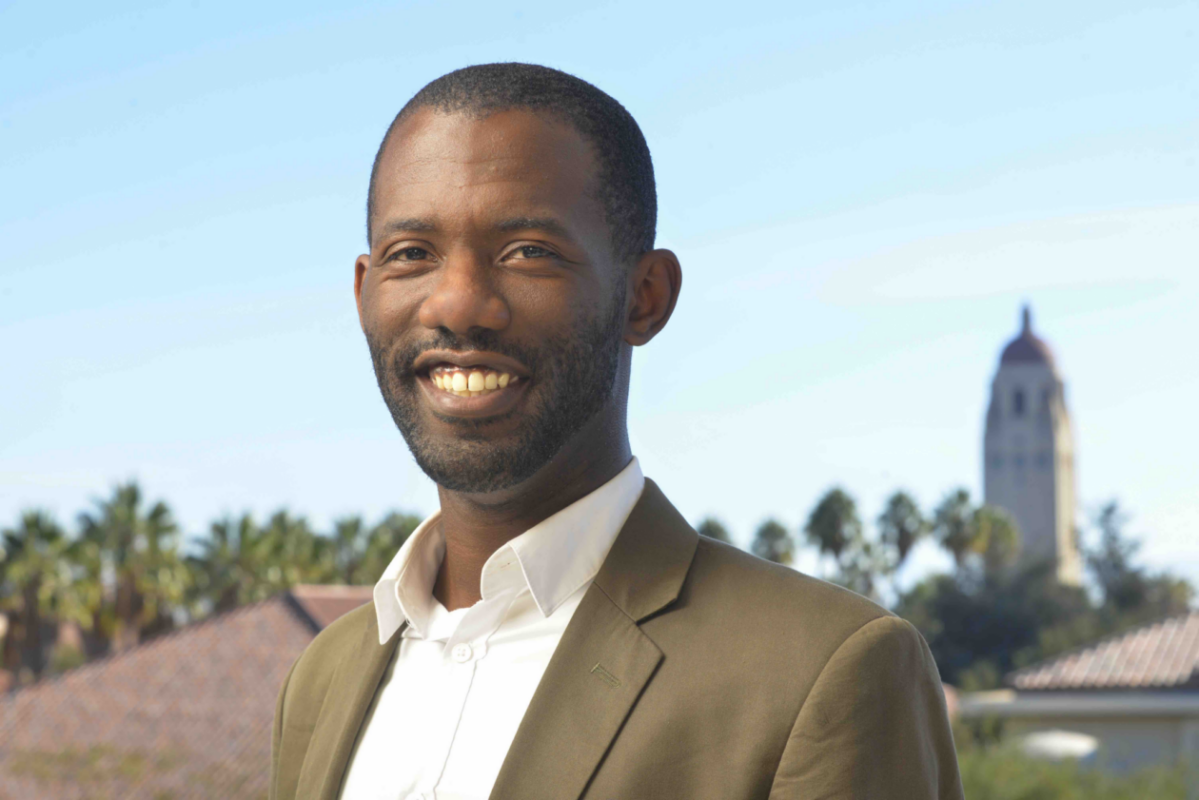 A man in a suit smiles outdoors with a clear blue sky. Palm trees and a tower are visible in the background, creating a calm, professional atmosphere.