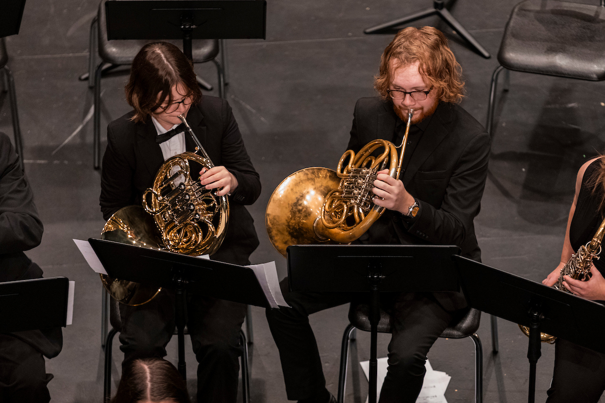 Two musicians in formal attire play French horns in an orchestra. They are seated on a stage with music stands, surrounded by a focused atmosphere.