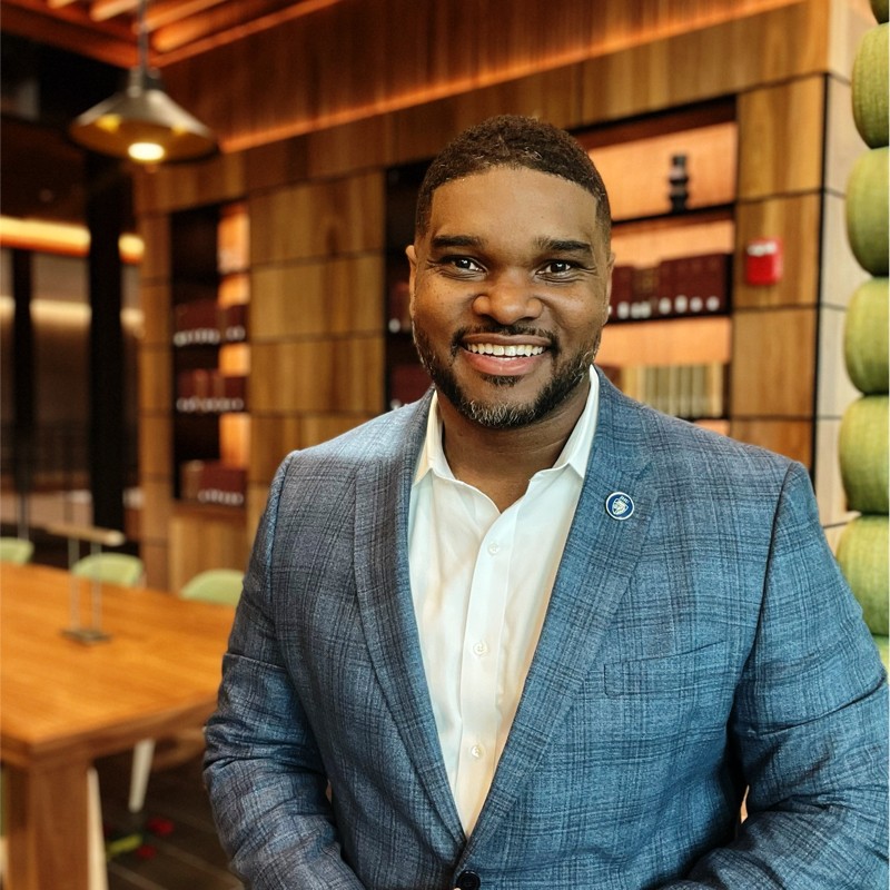 Smiling man in a blue blazer stands in a warmly lit, wood-paneled room with a modern design. The atmosphere is professional and welcoming.
