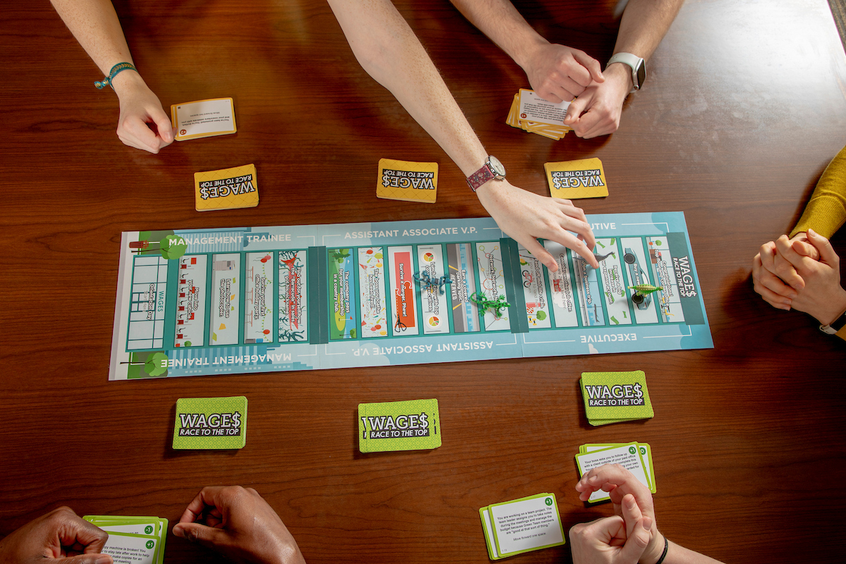 Hands of several people gathered around a table playing a board game called “WAGES: Race to the Top,” with cards and game pieces placed on the game board.