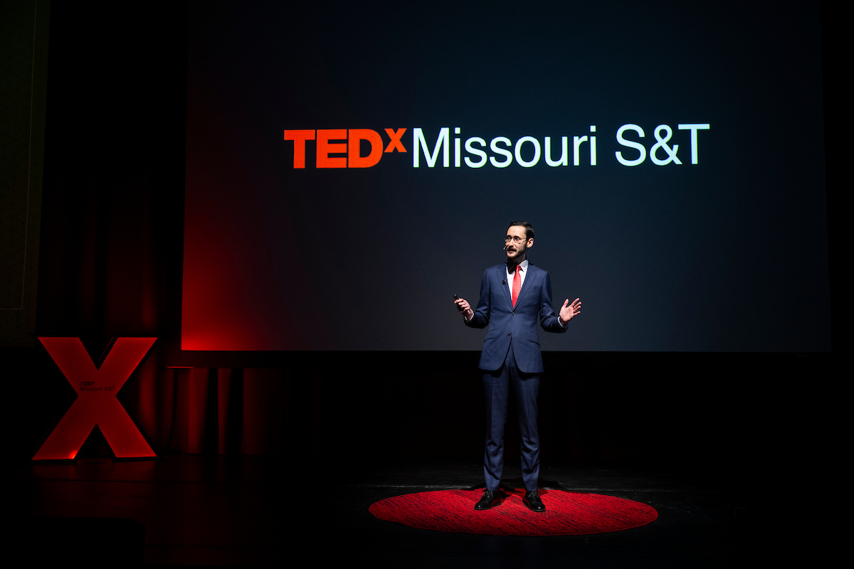 A man stands on the stage at Leach Theatre in front of a screen that reads TEDxMissouri S&T. He is speaking with his hands raised.