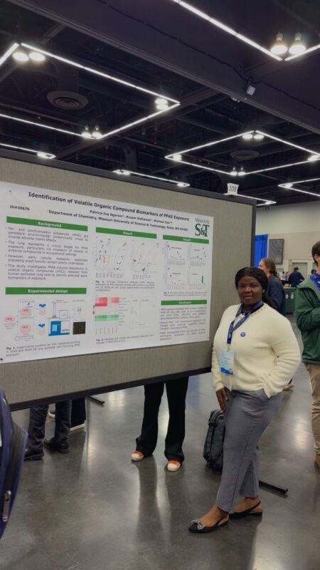 A woman stands near a board where her research poster is displayed.