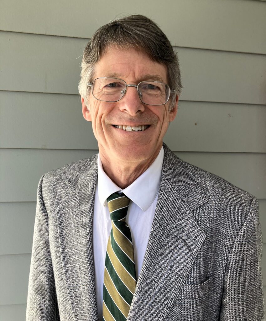 A man wearing glasses smiles for the camera wearing a grey jacket and tie.