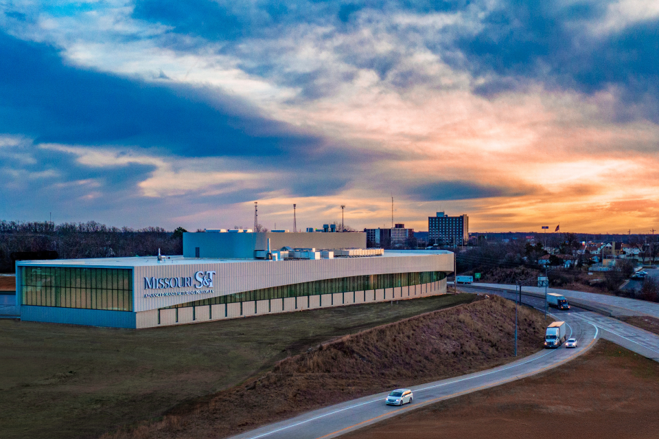 A modern building labeled "Missouri S&T Advanced Manufacturing Protoplex" is set against a vibrant sunset sky, with a highway and sparse traffic in the foreground.