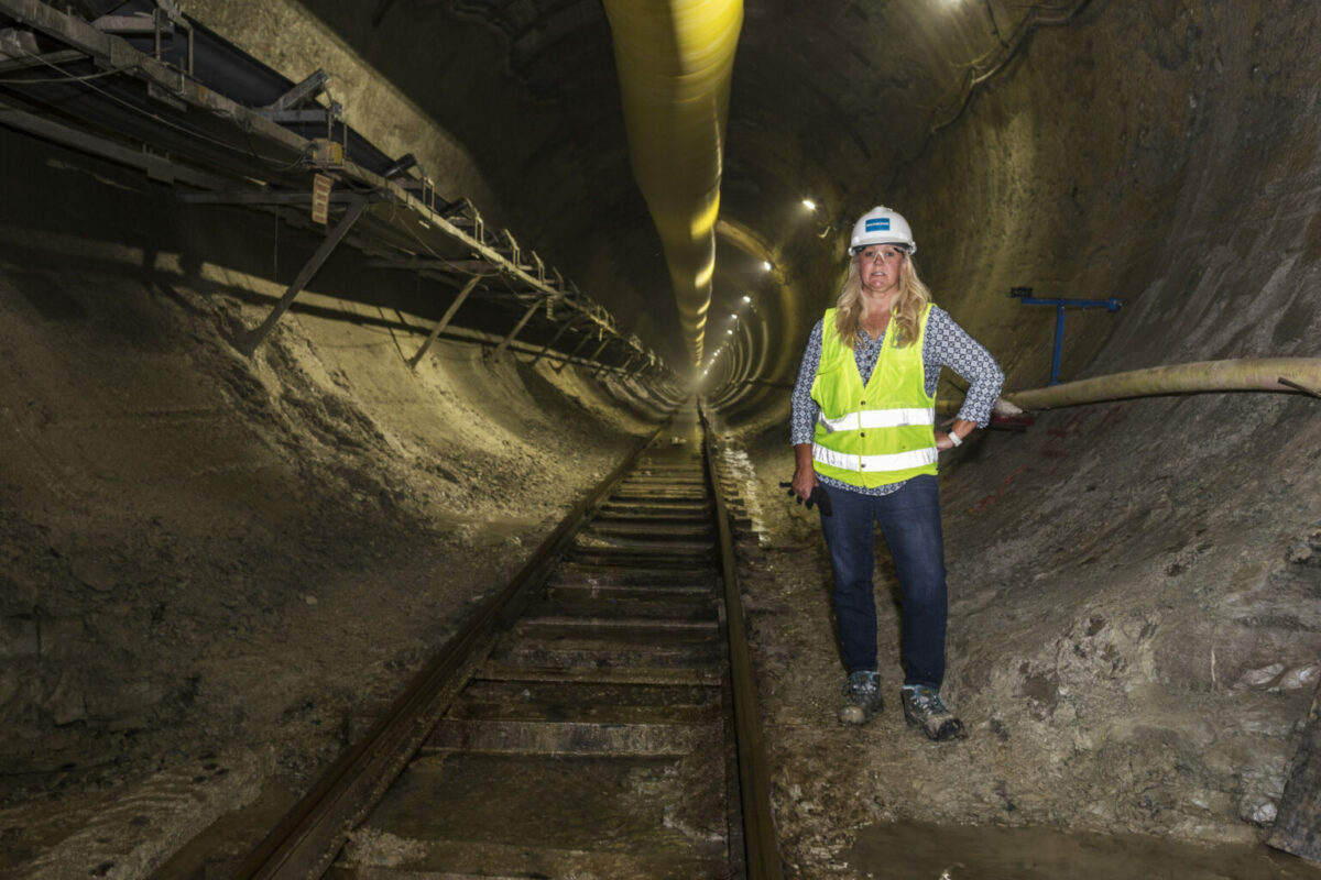 A person in safety gear stands beside rail tracks inside a wide underground tunnel lined with construction materials and lighting.