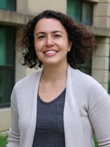 A photo of a woman wearing a light gray cardigan over a dark gray T-shirt in front of a brick building.