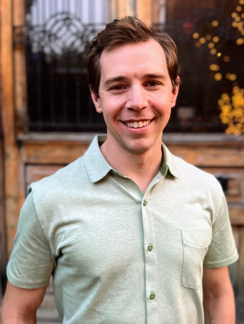A man wearing a light green button up collared shirt stands in front of an antique door.