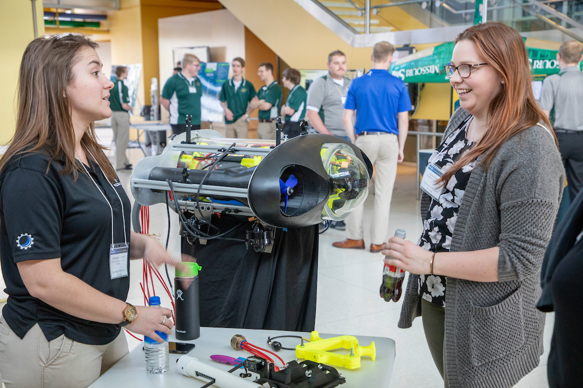 Two people stand at a table discussing a large cylindrical research device, with tools, equipment, and other attendees visible in the background.