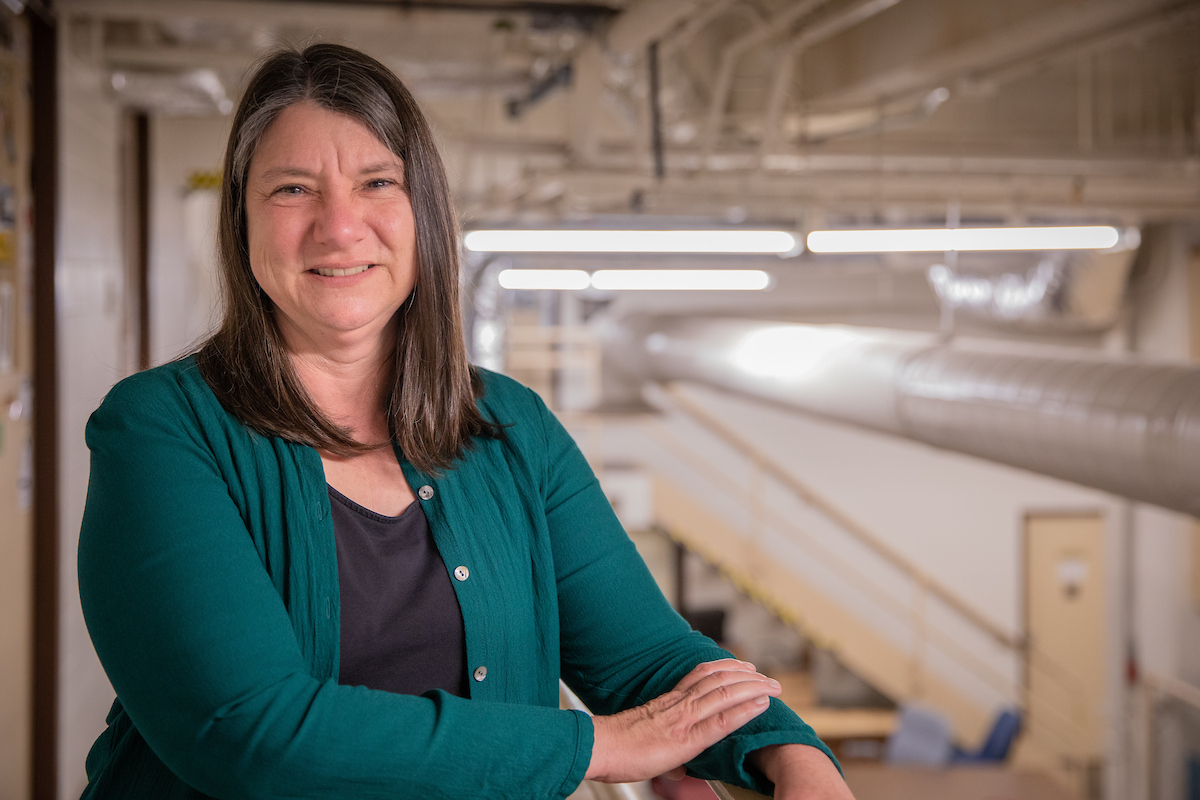 A woman wearing a teal cardigan and dark top smiles with her arms resting on a railing inside an industrial-style building. Exposed pipes, ductwork, and overhead lights extend into the background, creating a bright, modern workspace atmosphere.