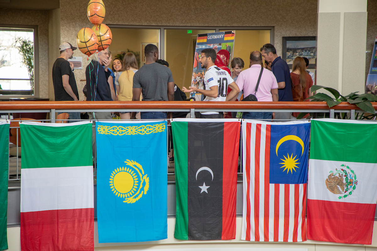 International flags hang along a railing inside a campus building while students gather and talk in the background during a multicultural event, with informational tables and decorations visible.