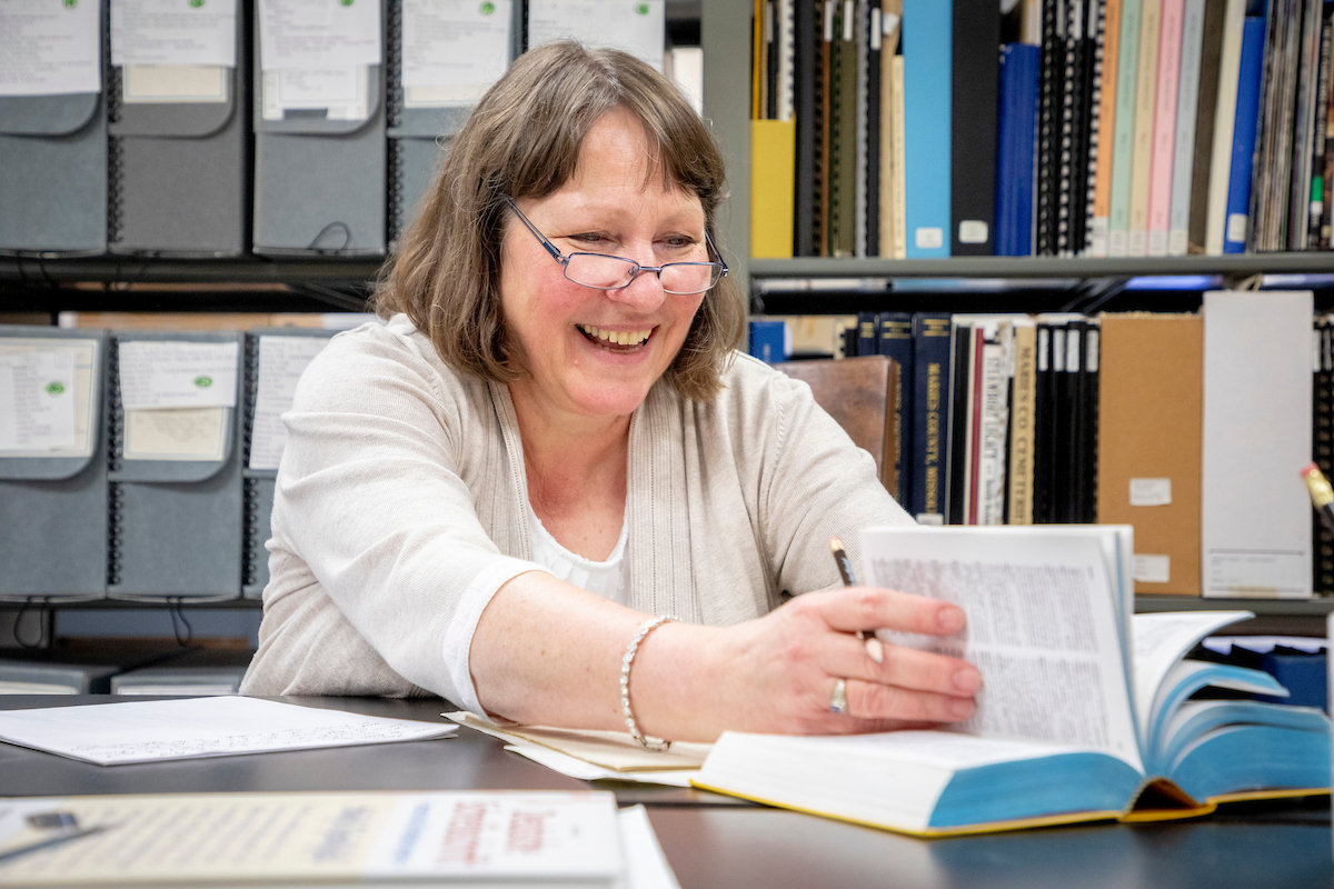 A woman wearing glasses looks through a book in a library with a cheerful smile.