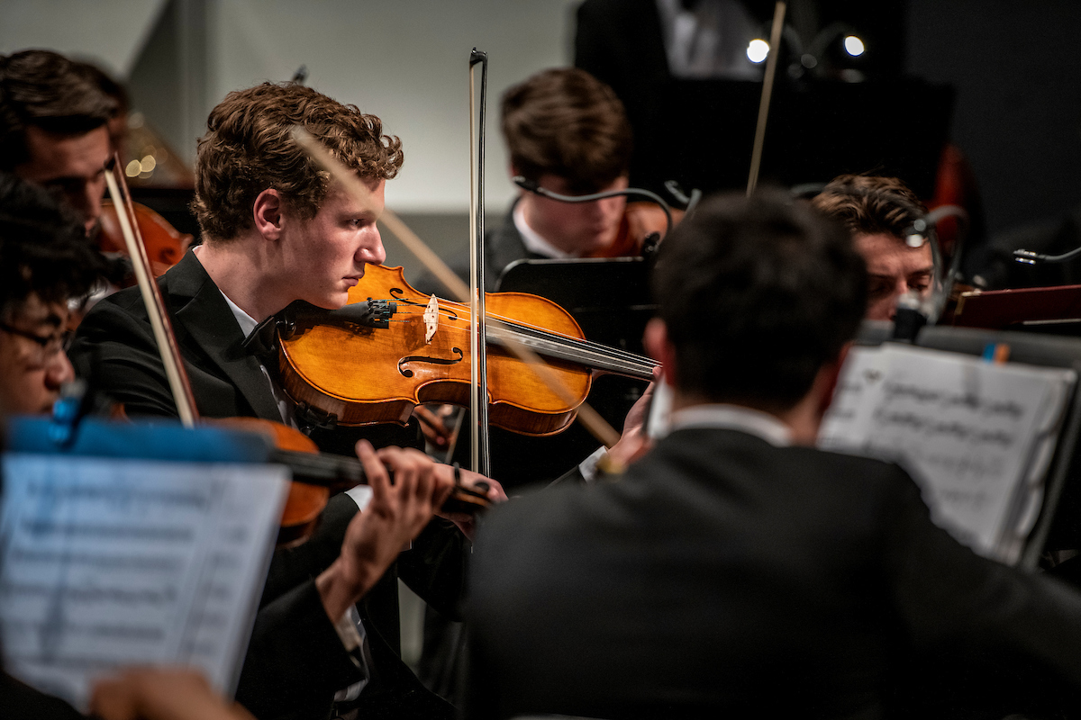 Orchestra musicians seated closely together while performing on stage, with several people playing string instruments such as violins and violas, reading from sheet music under soft stage lighting.