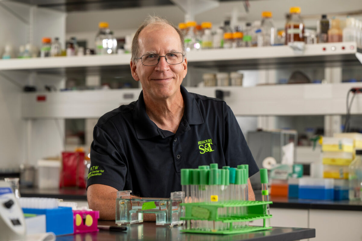 A person wearing a Missouri S&T polo stands at a lab bench with test tubes, pipette racks, and research equipment, with laboratory shelves in the background.
