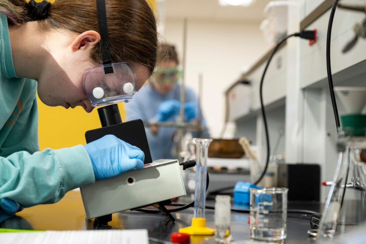 Two students wearing personal protective equipment work in a lab looking through microscopes.