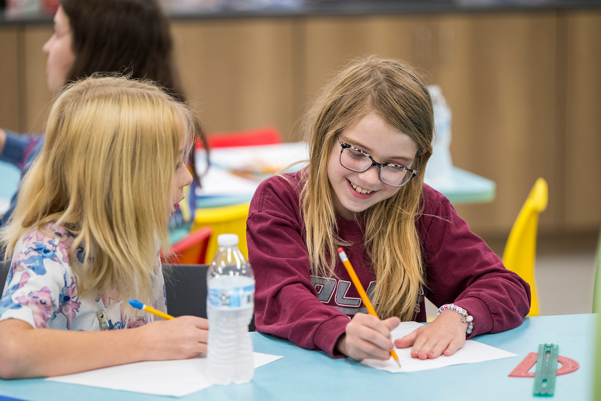 Two children sit at a table in a classroom or activity space, writing on paper with pencils. A water bottle, ruler, and craft supplies are on the table, with additional tables and chairs visible in the background.