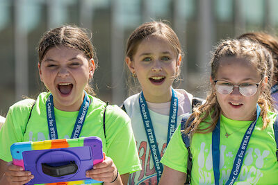 Three girls wearing blue lanyards and one holding a tablet look on in excitement as they discover something new at STEM Exploration Day.