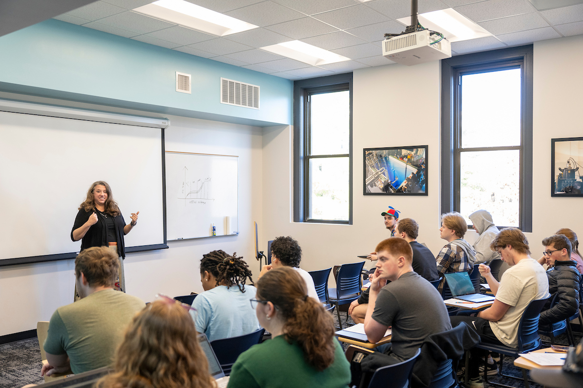 A woman stands in front of a white board in a classroom teaching several students seated at desks.