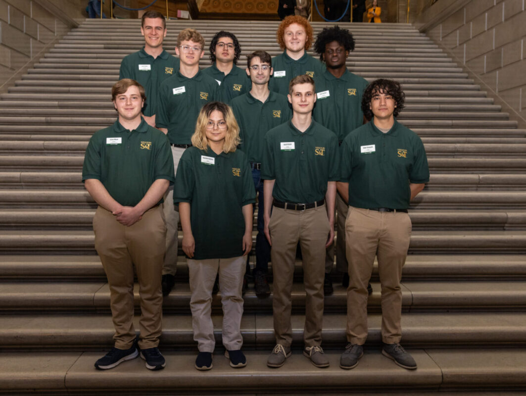 A group of twelve students wearing matching dark green polo shirts and khaki pants stand in three rows on a wide stone staircase inside a campus building, facing the camera and smiling.