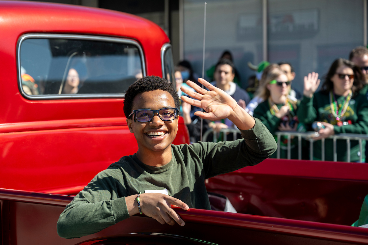 A smiling person wearing glasses waves from the back of a red vintage truck while spectators watch from behind barriers.