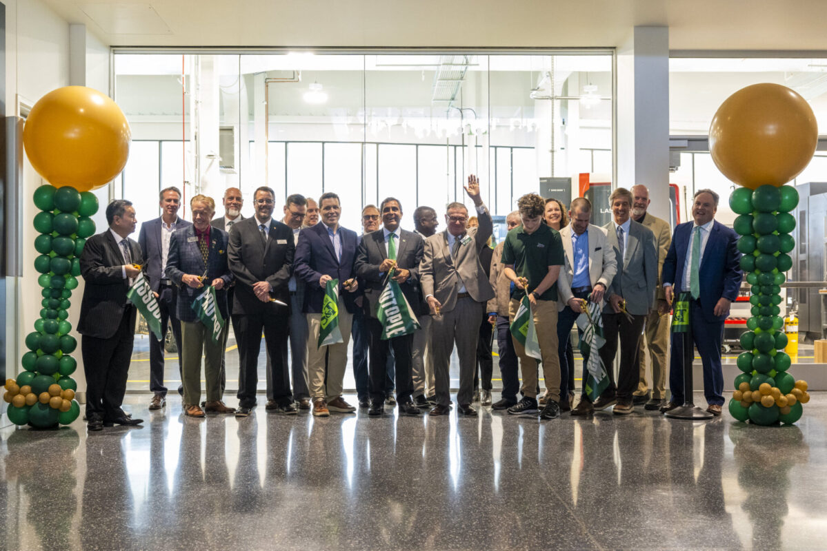 Group holding ceremonial ribbons inside a modern lobby, flanked by green and gold balloon columns.