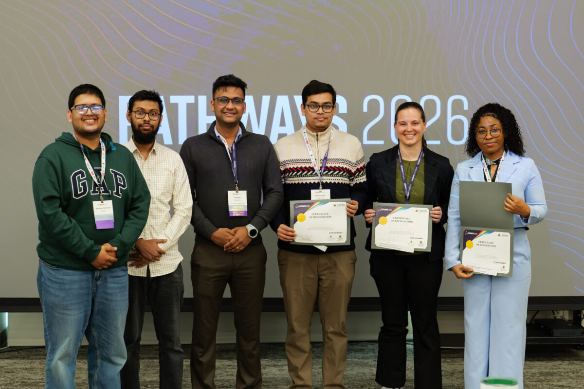 Seven people stand side by side on a stage in front of a projected screen reading “Pathway 2026,” with four individuals holding certificates. They are dressed in business casual attire, wearing conference badges, and smiling toward the camera.