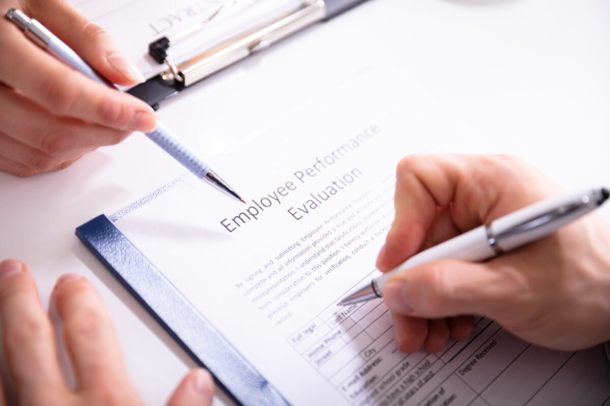 Two people hold pens while completing an employee performance evaluation form on a clipboard, with one person pointing to the document as the other writes, symbolizing workplace review and assessment.