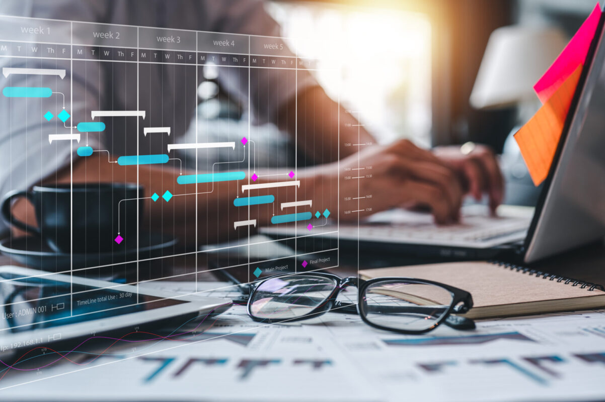 Person typing on a laptop at a desk with charts, glasses, and a digital project timeline graph overlay.