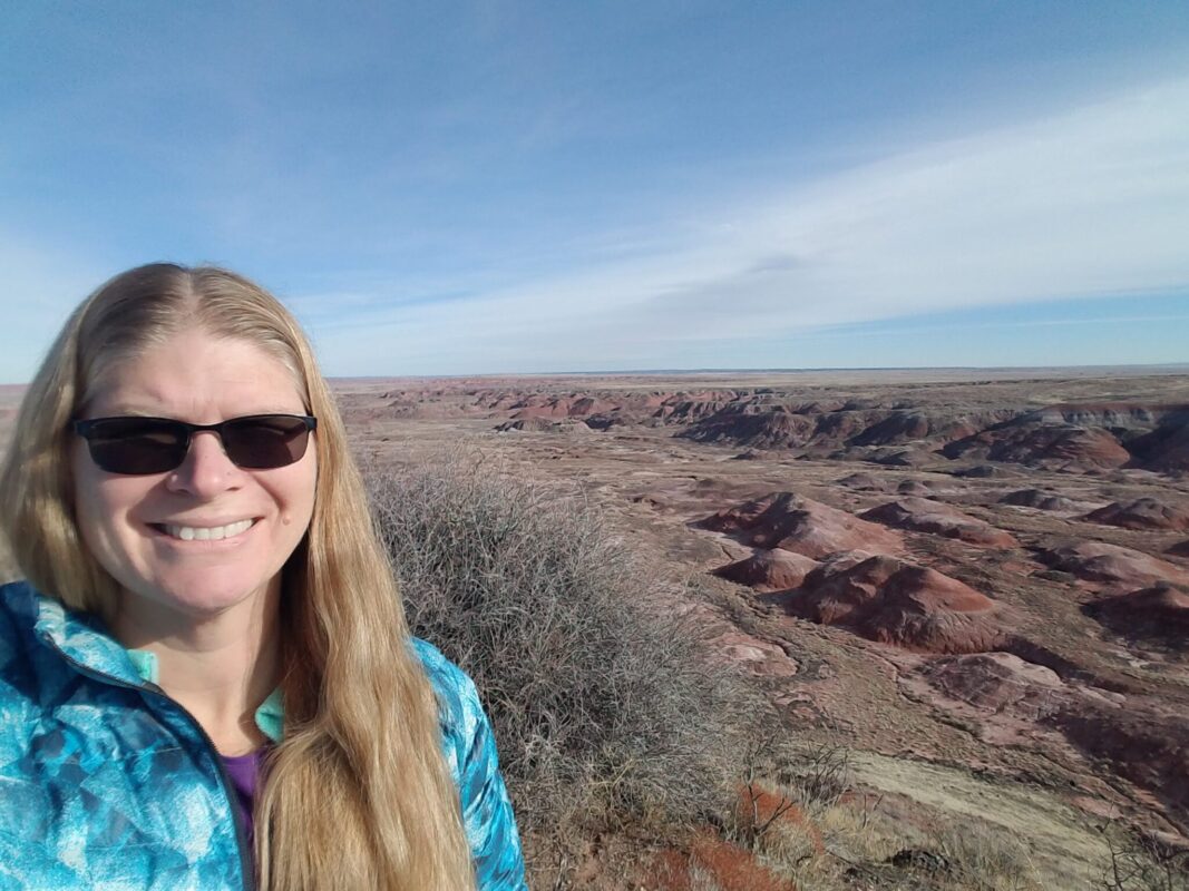 Woman with long blonde hair and sunglasses poses with the desert of Arizona in the background.