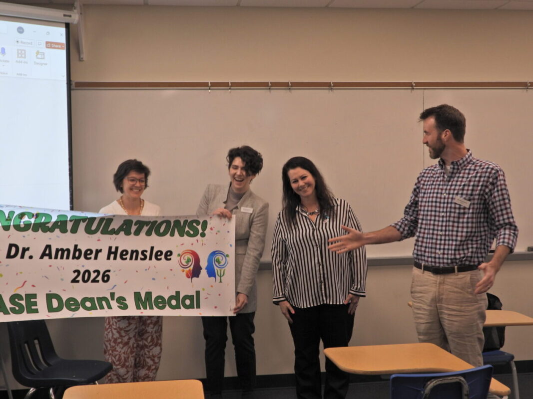 Four people stand in a classroom in front of a whiteboard, smiling and celebrating while two hold a colorful banner that reads “Congratulations! Dr. Amber Henslee – 2026 CASE Dean’s Medal.” Desks and chairs are visible in the foreground as one person gestures toward the banner.
