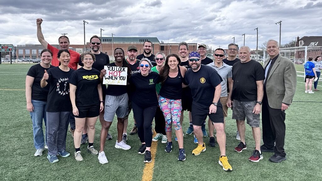 Members of the faculty kickball team stand together on an outdoor athletic field after the game, wearing athletic clothing and posing in a group under a cloudy sky.