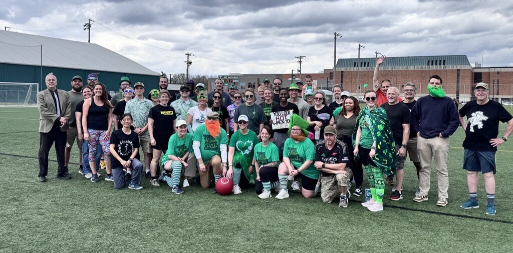 Faculty and staff participants pose together on an outdoor athletic field after the Faculty vs. Staff Kickball Game, with both teams standing and kneeling in rows under a cloudy sky.