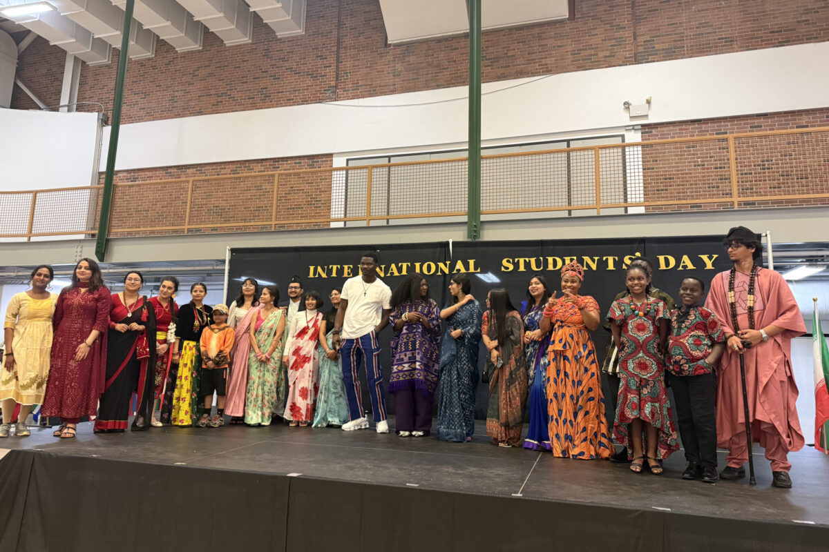 Group of people in colorful traditional outfits standing on a stage beneath an “International Students Day” banner.