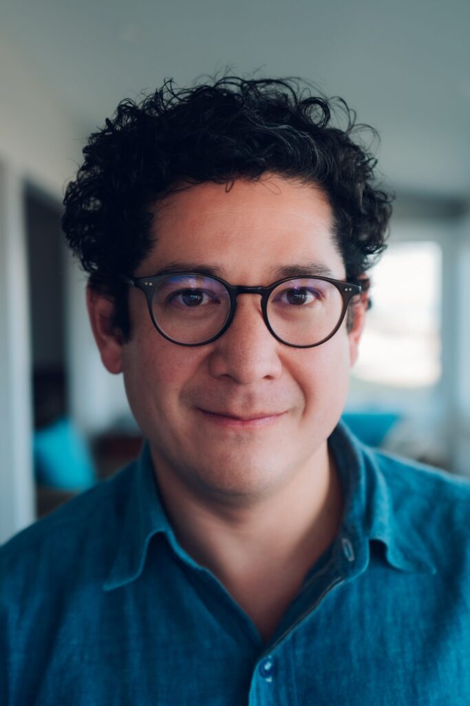 Close-up portrait of a person with short, curly dark hair and round glasses, wearing a blue button-up shirt, indoors with a softly blurred background and natural light.