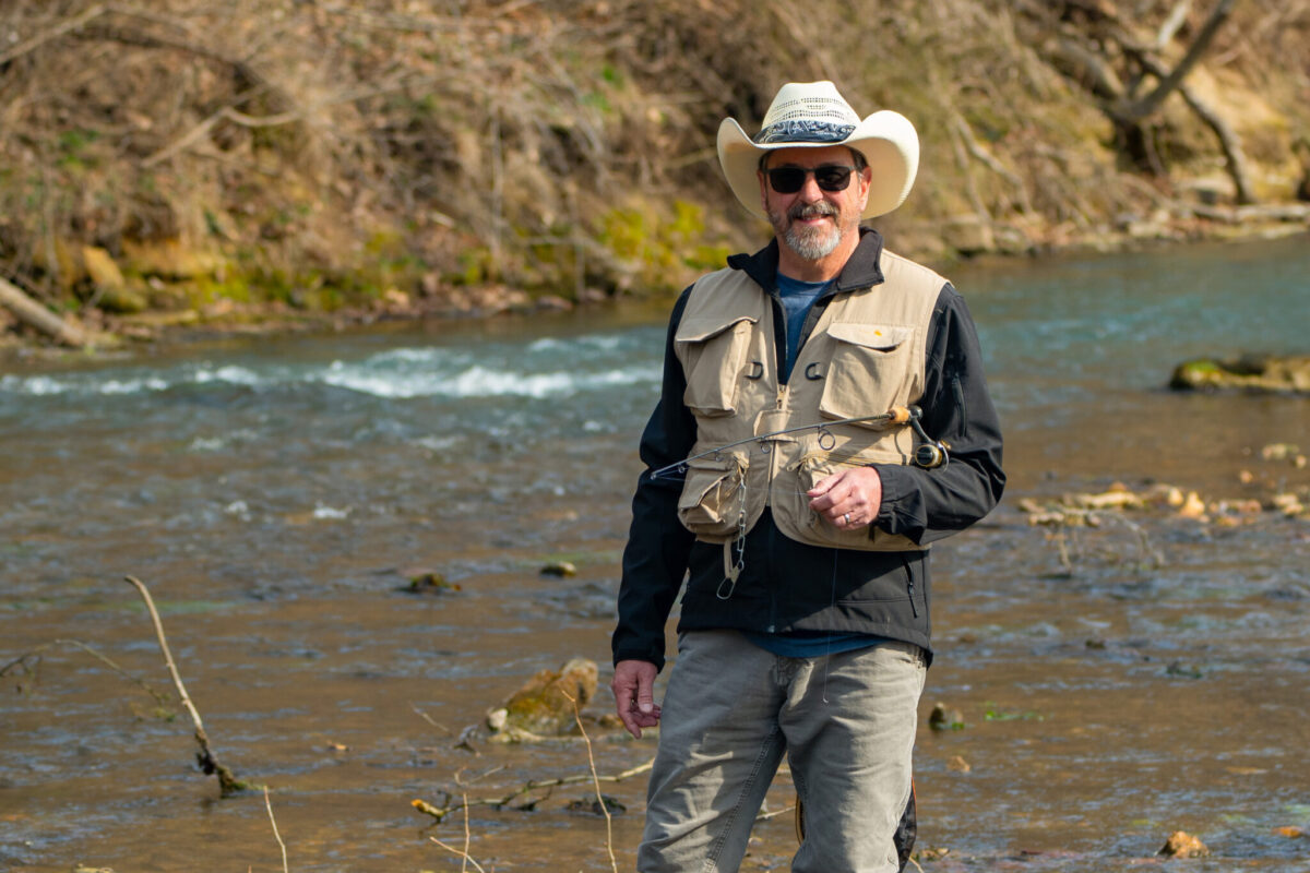 Person wearing a cowboy hat and fishing vest stands in a shallow creek holding a fishing rod, with trees and rocks in the background.